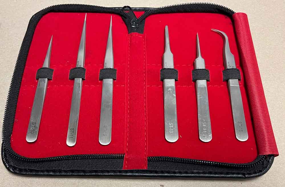 Set of six stainless tweezers with varied tip types  in a red case with black straps on a beige background